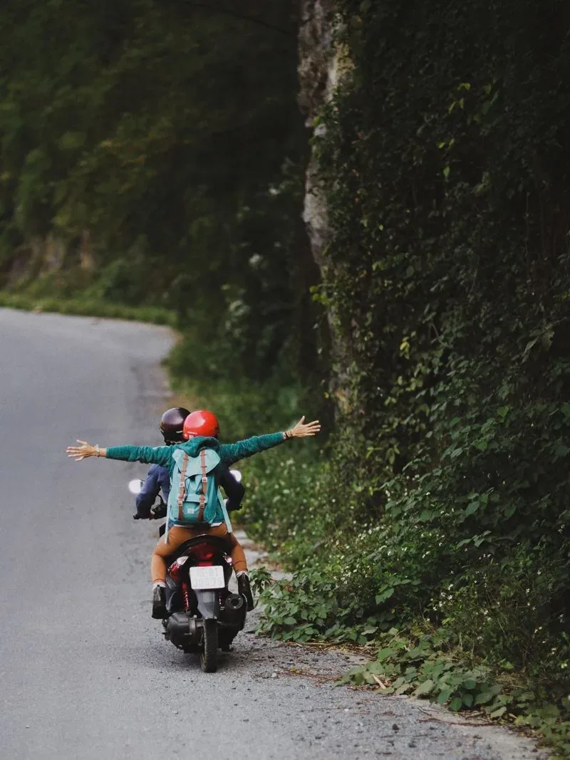 Two riders with arms outstretched on a moped down a forest road