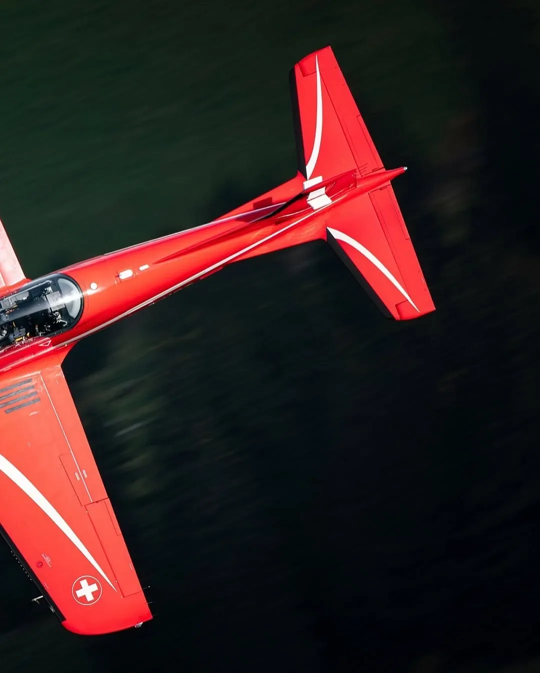 A red Swiss aerobatic aircraft banking hard, seen from directly above over dark forest