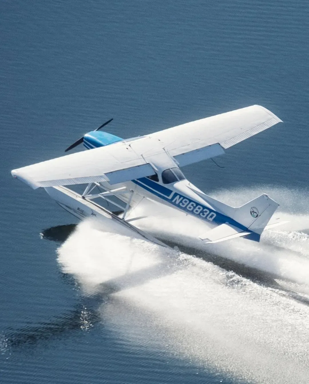 A white and blue floatplane carving a turn on the water, throwing up a huge spray