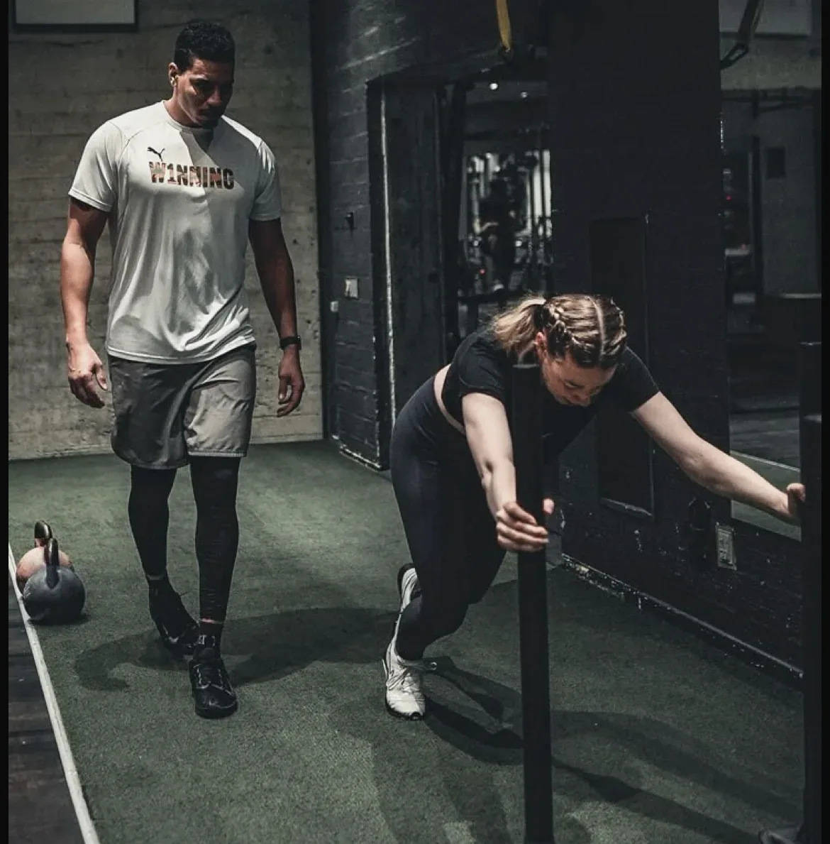 A coach walking a client through a sled and kettlebell session in a dark, concrete gym