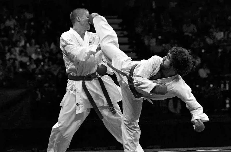 Two karate competitors mid-kumite, a high kick landing, shot in black and white